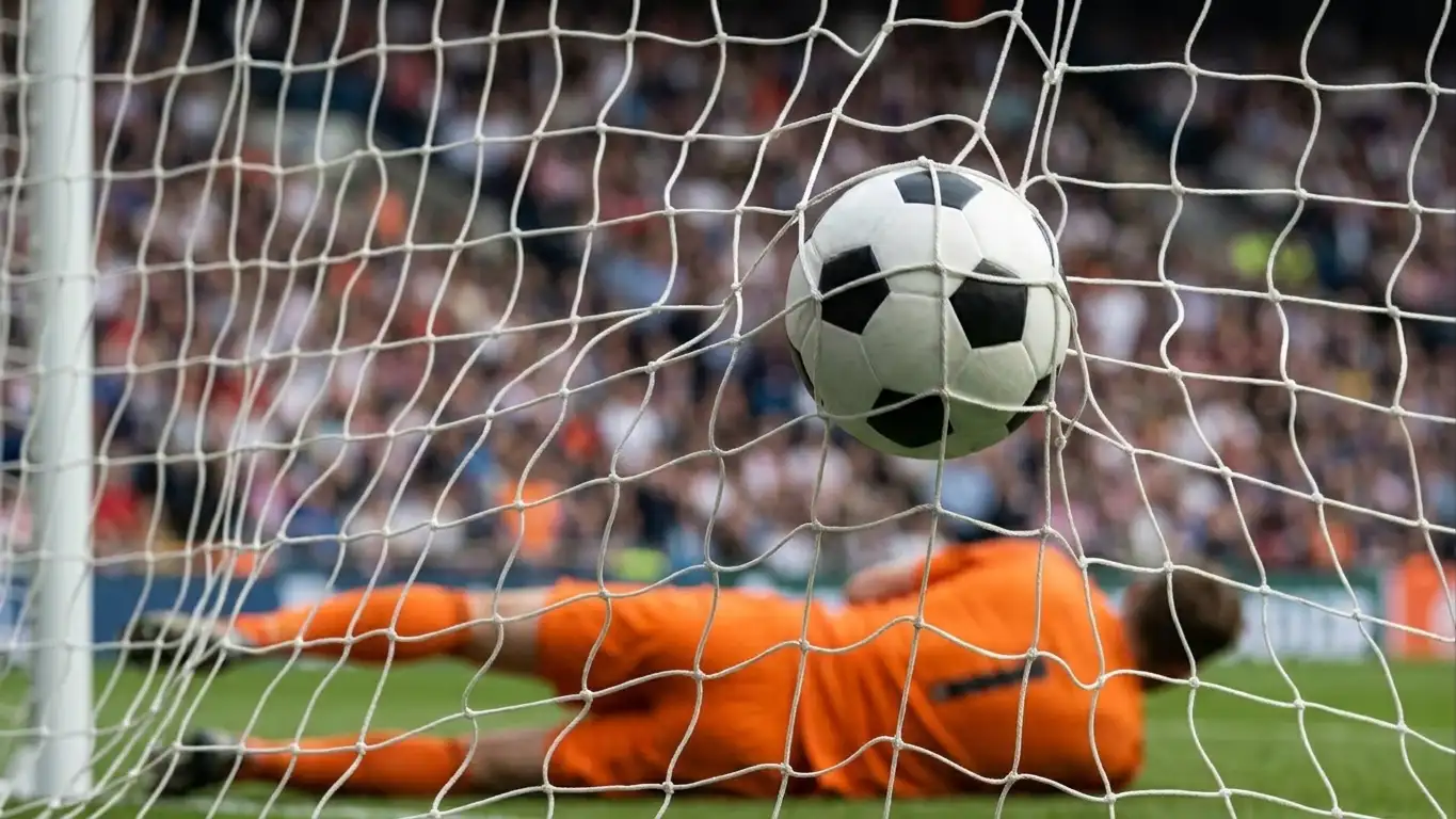 A football hitting the back of a net, with the goalkeeper on the ground and crowd blurred behind